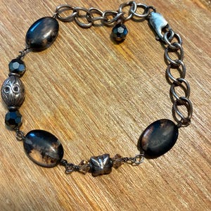 May include: A bracelet featuring dark brown and black oval stones, faceted black beads, and silver-toned metal spacers. The design includes a silver-toned chain link section with a lobster claw clasp and a small dangling bead charm attached to the chain.