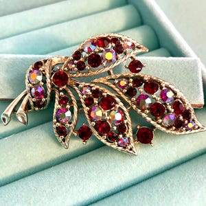 May include: A gold-tone metal brooch shaped like a cluster of leaves, featuring a textured finish and embedded with numerous round, faceted red crystals and iridescent stones that catch the light with various colors throughout the leaf design.