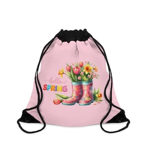 Può includere: Una borsa rosa con coulisse e chiusura con coulisse nera. La borsa presenta un'illustrazione ad acquerello di stivali di gomma rosa pieni di tulipani e narcisi. Le parole "hello SPRING" sono stampate sulla borsa.