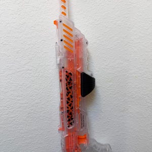May include: A clear plastic toy blaster with bright orange accents on the barrel, top vents, and internal components. The side features a black handle grip and a decorative panel with a repeating black bat pattern. Text reads DO NOT SHOOT IN FACE OR EYES.
