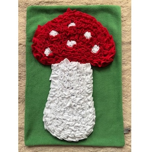 Könnte beinhalten: Ein handgefertigtes Kissen in Pilzform. Es hat einen roten, gerüschten Hut mit weißen Punkten, einen weißen, gerüschten Stiel und einen grünen Hintergrund. Das Kissen ist aus weichem Stoff gefertigt.