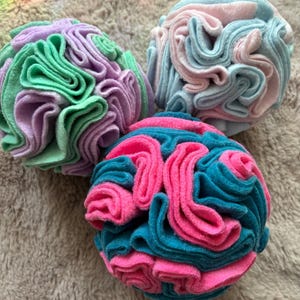 Op de afbeelding: Drie decoratieve vilten ballen in groen, paars, roze en blauwgroen. De ballen hebben een gelaagd, gerimpeld ontwerp. De vilten ballen zijn gerangschikt op een zacht, neutraal getint oppervlak. De ballen zijn decoratief.