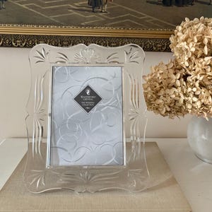 May include: A clear crystal picture frame with a scalloped edge and etched fan patterns. The frame features a central heart motif at the top and contains a display card with the text Waterford Crystal The Wedding Collection in a diamond shape.