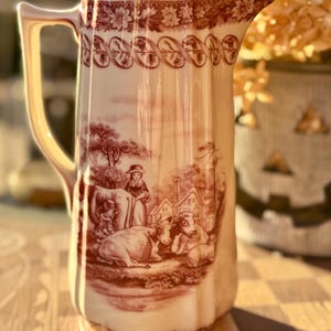 May include: A cream-colored ceramic pitcher featuring a deep red transferware pattern. The design depicts a rural scene with a person, cows, and buildings. The rim is decorated with a floral and ribbon border, and the handle is curved.