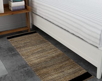 May include: A rectangular, woven jute rug with a natural tan and black color scheme. The rug features a black border at each end and is placed on a dark gray floor next to a bed with white bedding.