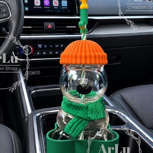 May include: A clear glass tumbler shaped like a snowman, featuring a bright orange ribbed silicone hat, a green knitted scarf, and a green silicone base. A striped straw passes through the center, topped with a small yellow flame-shaped topper.