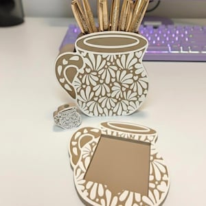 May include: A decorative desk set featuring a pen holder, coaster, and small trinket. The pen holder and coaster are shaped like mugs with a brown and white floral design. Gold pens with crystal tops are in the pen holder.