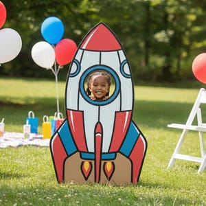 May include: A cardboard rocket ship cutout with a child's face visible in the porthole. The rocket is red, white, and blue, with flame details. Balloons in red, white, and blue are in the background, along with a picnic setup and white folding chairs.