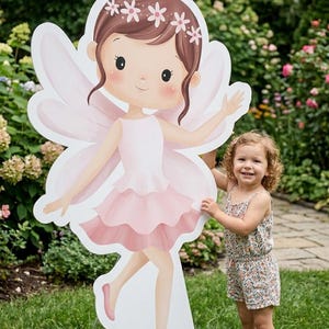 May include: A large, freestanding fairy cutout with a pink dress, wings, and floral headpiece. A young child in a floral romper stands next to the fairy cutout on a grassy lawn, smiling. The background features green foliage and a pathway.