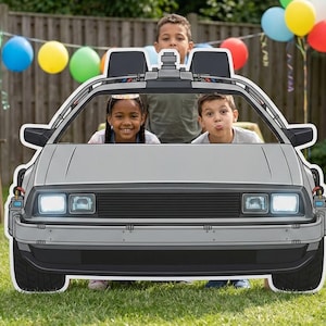 May include: A gray cardboard cutout of a DeLorean car with three children posing inside. The car has black details, including the wheels, trim, and roof accents. The background includes colorful balloons and a wooden fence.