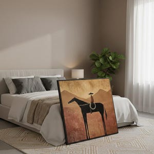May include: Framed art print featuring a minimalist illustration of a cowboy on a horse. The artwork has a black frame and depicts a rider in a hat on a black horse against a backdrop of brown and tan mountains and a red-brown field.