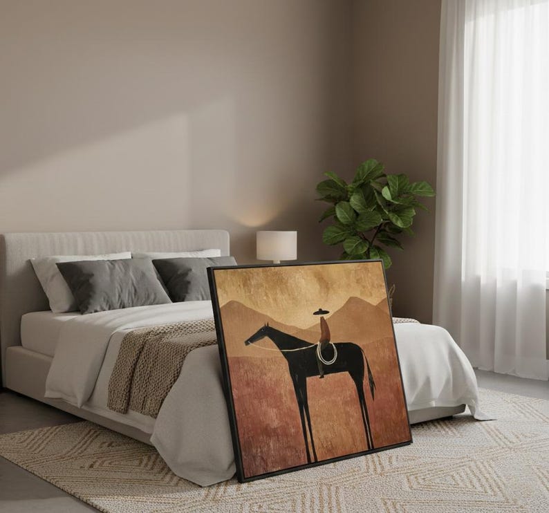 May include: Framed art print featuring a minimalist illustration of a cowboy on a horse. The artwork has a black frame and depicts a rider in a hat on a black horse against a backdrop of brown and tan mountains and a red-brown field.