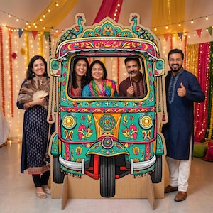 May include: A colorful cardboard cutout of a decorated auto-rickshaw, with a peacock design on top. Several people pose behind and beside the cutout. The auto-rickshaw is painted in shades of teal, pink, yellow, and orange, with floral patterns.