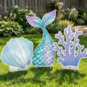 Könnte beinhalten: Drei Gartendekorationen: eine Muschel, eine Meerjungfrauenflosse und Korallen. Die Muschel ist hellblau, die Meerjungfrauenflosse ist türkis und rosa, und die Korallen sind lila. Alle drei Dekorationen stehen auf einem grünen Rasen.