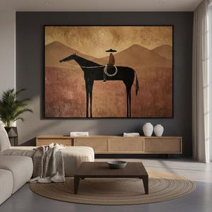 May include: Large framed artwork depicting a silhouetted rider on horseback. The artwork features a black horse and rider against a backdrop of brown mountains and a textured field. The piece is displayed in a modern living room setting.