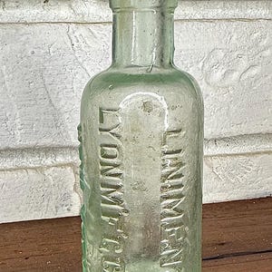 May include: An antique, light green glass bottle with the embossed text "LYDN M'F'G CO" and "LINIMENT". The bottle has a rounded body and a narrow neck. The bottle is displayed against a white brick wall.
