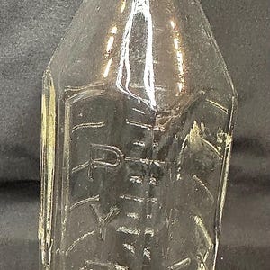 May include: A clear, vintage glass bottle with a rectangular shape and a narrow neck. The bottle has embossed text and measurement markings. The bottle is empty and appears to be an antique milk bottle.