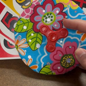 May include: A red fidget spinner rests on a floral patterned coaster with a blue background. The coaster has pink and red flowers, green leaves, and silver embellishments. Uno cards are in the background.