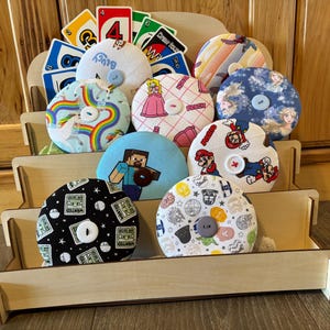 May include: A wooden display stand holds a collection of colorful, round fabric items with various designs, including characters from Mario, Minecraft, and Disney's Frozen. Some items have buttons. Uno cards are in the background.