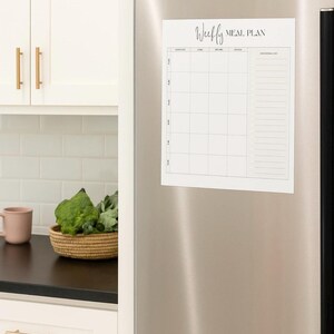 Puede incluir: Un calendario magnético blanco de planificación semanal de comidas con un diseño de cuadrícula y una sección de lista de compras, adherido a un refrigerador de acero inoxidable. Las palabras "Weekly Meal Plan" están escritas en la parte superior en cursiva.