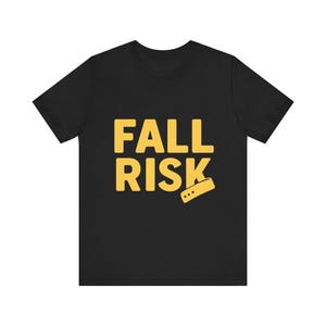 May include: Black t-shirt with the words "FALL RISK" in yellow block letters. A yellow rectangle with three dots is below the word "RISK". The shirt is made of a soft material.