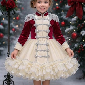 May include: A young girl wearing a festive dress with a burgundy velvet bodice and cream-colored skirt. The dress features silver trim, decorative buttons, and a ruffled skirt. The image includes the text "Flower Closet".