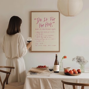 Puede incluir: Escena de comedor con una obra de arte enmarcada que dice "Do It For the Plot". Una persona sostiene una copa de vino tinto. La mesa está puesta con una botella de vino, tomates, platos y flores.