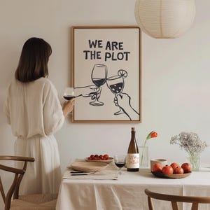 May include: A framed print with the words "WE ARE THE PLOT" and a graphic of two hands toasting wine glasses. A woman in a cream dress holds a glass of red wine. A table is set with a bottle of wine, fruit, and flowers.