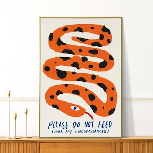 Könnte beinhalten: Ein gerahmter Kunstdruck mit einer stilisierten orangefarbenen Schlange mit schwarzen Punkten auf cremefarbenem Hintergrund. Die Schlange hat ein blaues Auge und eine rote Zunge. Der Text "PLEASE DO NOT FEED (UNDER ANY CIRCUMSTANCES)" ist blau geschrieben.