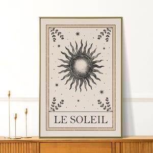 May include: A framed print featuring a detailed illustration of the sun with radiating lines, surrounded by stars and botanical accents. The text "LE SOLEIL" is at the bottom. The artwork has a vintage aesthetic with a beige and black color palette.