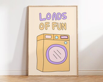 Op de afbeelding: Een ingelijste print met een cartoon illustratie van een wasmachine in geel, paars en blauw. De woorden "LOADS OF FUN" staan boven de machine in paarse en oranje bubbelletters. Het kunstwerk wordt gepresenteerd op een lichtbeige achtergrond.