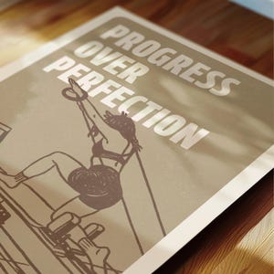 May include: A poster with the text "PROGRESS OVER PERFECTION" in white letters. The poster features a line drawing of a person exercising on a Pilates machine. The color palette is neutral, with shades of beige and brown.
