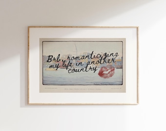 Pode incluir: Impressão emoldurada com o texto "Brb, romanticizing my life in another country" e uma marca de beijo vermelha. A imagem apresenta uma cena à beira-mar. A moldura é de cor clara.