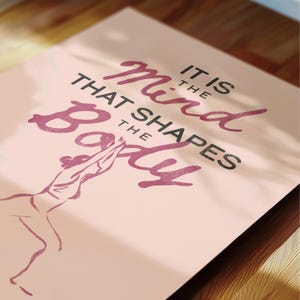 May include: A pink poster with the quote "It is the mind that shapes the body" in a mix of dark pink and black fonts. A line drawing of a person in a yoga pose is at the bottom. The poster is on a wooden surface.