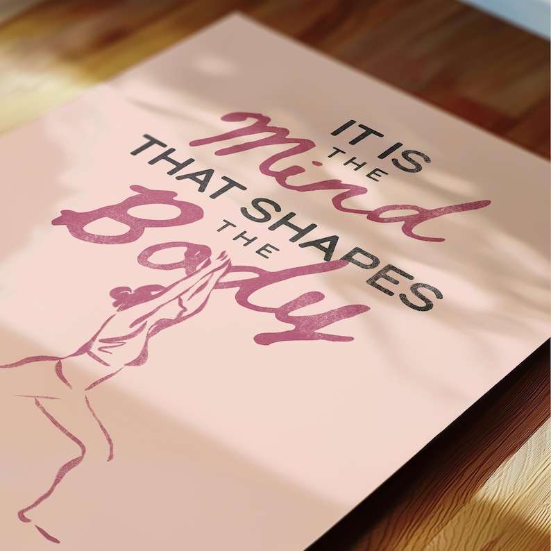 May include: A pink poster with the quote "It is the mind that shapes the body" in a mix of dark pink and black fonts. A line drawing of a person in a yoga pose is at the bottom. The poster is on a wooden surface.