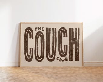 May include: A framed print with the words "THE COUCH CLUB" in a brown, textured font against a beige background. The print is in a light brown wooden frame and leans against a white wall on a wooden floor.