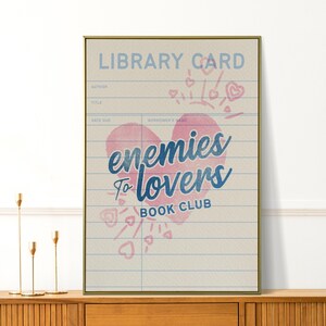 Puede incluir: Impresión enmarcada con borde dorado. El diseño es una tarjeta de biblioteca vintage con el texto "LIBRARY CARD" y "enemies to lovers BOOK CLUB" en azul y rosa. El diseño está decorado con corazones y flechas.