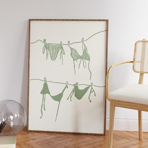 Può includere: Una stampa artistica incorniciata con un'illustrazione minimalista di quattro bikini verdi appesi a un filo. L'opera d'arte è ambientata su uno sfondo color crema, con un'estetica semplice e disegnata a linea. La cornice è di colore marrone caldo.
