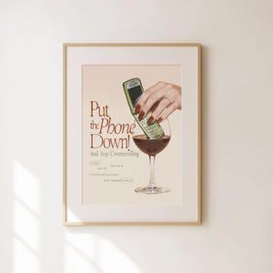May include: A framed wall art print featuring a green flip phone submerged in a glass of red wine. The text reads: Put the Phone Down! And Stop Doomscrolling. The design includes additional small text and a hand with dark red nail polish.