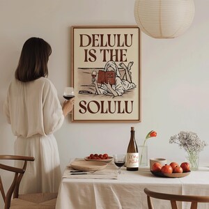 May include: A rectangular wall art print in a wooden frame featuring a line drawing of a person in a robe lying down, reading a book, and holding a wine glass. The text reads DELULU IS THE SOLULU in bold brown letters against a cream background.