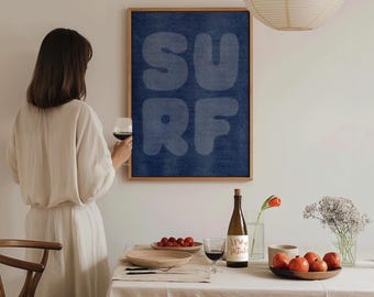 May include: A framed wall art print with the word "SURF" in large, light blue letters against a dark blue textured background. The print is in a wooden frame. A table is set with a white tablecloth, wine, fruit, and flowers.