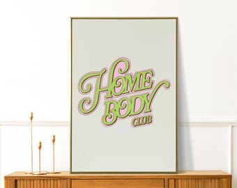 May include: A framed art print with the words "Home Body Club" in a retro font. The text is green with a pink and brown outline, set against a light green background. The frame is gold, and the print is displayed on a wooden cabinet.