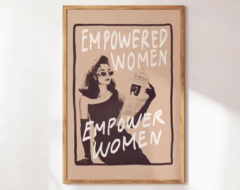 May include: A framed poster with the words "EMPOWERED WOMEN EMPOWER WOMEN" in white, handwritten-style font. The image features a woman in sunglasses and a dress, holding a newspaper. The poster has a tan background and a wooden frame.