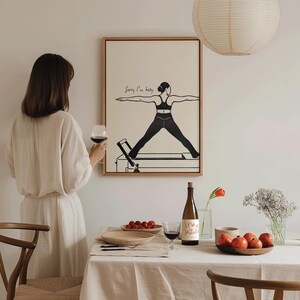 May include: A framed artwork with a minimalist illustration of a person doing Pilates, with the text "Sorry I'm busy." A woman in a cream-coloured dress holds a glass of red wine. A table is set with fruit, wine, and a vase of flowers.