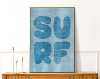 May include: A framed wall art print with the word "SURF" in large, blue, bubble letters. The letters are on a light blue textured background, and the frame is gold. The artwork is displayed on a wooden cabinet.