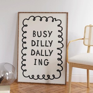 May include: A framed art print with the words "BUSY DILLY DALLY ING" in black, outlined by a black squiggly border, on a cream background. The frame is a warm brown color. A chair with a gold frame and woven back is to the right.