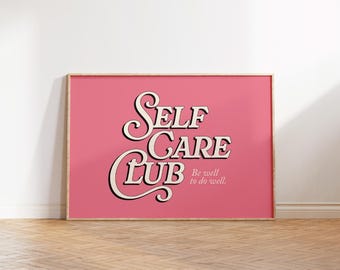 May include: A framed pink poster with the words "SELF CARE CLUB" in a vintage font. Below the words is the phrase "Be well to do well." The frame is a light wood color, and the background is a white wall and wood floor.