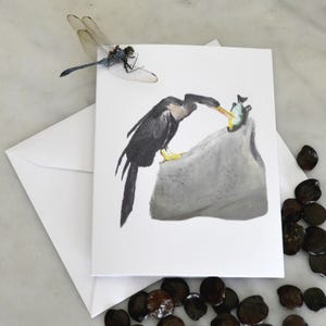 May include: A white greeting card featuring a watercolor illustration of a black bird with yellow feet eating a fish. A dragonfly rests on the card. An envelope and scattered seeds are also visible.