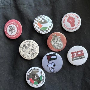 May include: Eight circular badges with diverse designs and text. The badges display the Palestinian flag, Snoopy with a Palestinian flag, and slogans such as "Free Palestine" and "Occupation is a Crime". The badges are on a black surface.