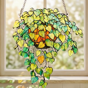 Puede incluir: Un atrapasoles de vidrieras con forma de planta colgante con enredaderas en cascada. Las hojas están hechas de varios tonos de vidrio verde y amarillo, mientras que la maceta es naranja. Cuelga de una cadena de metal plateado ante una ventana.